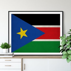 Puede incluir: Una impresión enmarcada de la bandera de Sudán del Sur. La bandera es negra, roja, verde y azul con una estrella amarilla de cinco puntas en el triángulo azul.