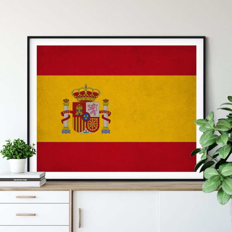 May include: Framed print of the Spanish flag, featuring a red and yellow design with the Spanish coat of arms. The flag is rectangular and has a distressed, vintage appearance. The frame is black, and the print is displayed on a white cabinet.
