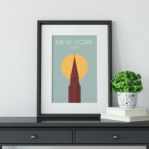 May include: A black framed print with a minimalist design featuring a red skyscraper against a light blue background with a yellow sun. The text "NEW YORK USA" is at the top of the print.