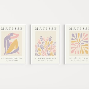 May include: Three minimalist art prints featuring the work of Henri Matisse. The prints are in a white frame and feature a light beige background with a pink, yellow, and purple abstract design. The text "MATISSE" is at the top of each print. The text below the design on each print reads "GALERIE D'EXPOSITION Papier D&eacute;coup&eacute;", "AIX-EN-PROVENCE Pavillon de Vendome", and "MUS&Eacute;E D'ORSAY Rue de la L&eacute;gion d'honneur".