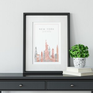 May include: A black framed print with a watercolor illustration of the New York City skyline in shades of pink and gray. The text "NEW YORK" and "UNITED STATES" is printed at the top of the print.