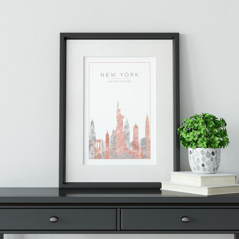 May include: A black framed print with a watercolor illustration of the New York City skyline in shades of pink and gray. The text "NEW YORK" and "UNITED STATES" is printed at the top of the print.