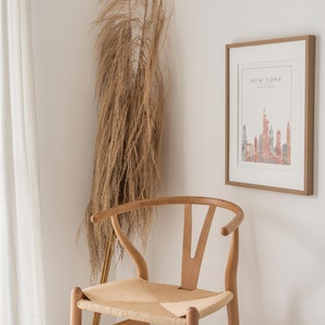 May include: A framed print with a watercolor illustration of the New York City skyline in shades of pink and gray. The print is hanging on a white wall next to a wooden chair with a woven seat.