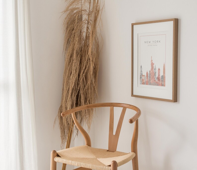 May include: A framed print with a watercolor illustration of the New York City skyline in shades of pink and gray. The print is hanging on a white wall next to a wooden chair with a woven seat.