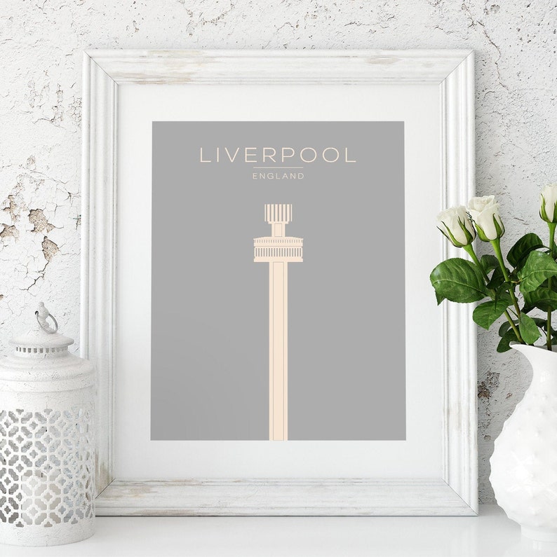 May include: A framed print with a gray background and a white outline. The print features a simple illustration of a tall tower in a light peach color. The text "LIVERPOOL" is at the top of the print in a white font, and the text "ENGLAND" is below it in a smaller white font.