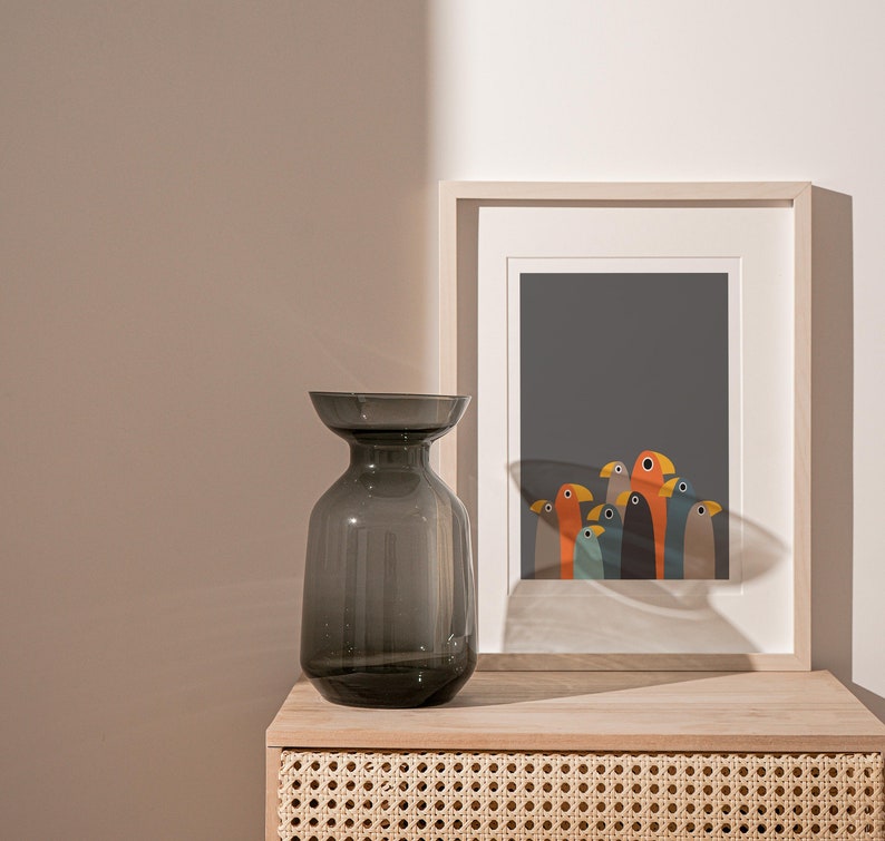 K&ouml;nnte beinhalten: Eine dunkelgraue Glasvase mit einer breiten &Ouml;ffnung steht auf einem hellen Holztisch mit einer Schublade aus geflochtenem Rattan. Hinter der Vase befindet sich ein gerahmter Druck einer Gruppe bunter V&ouml;gel auf dunkelgrauem Hintergrund.