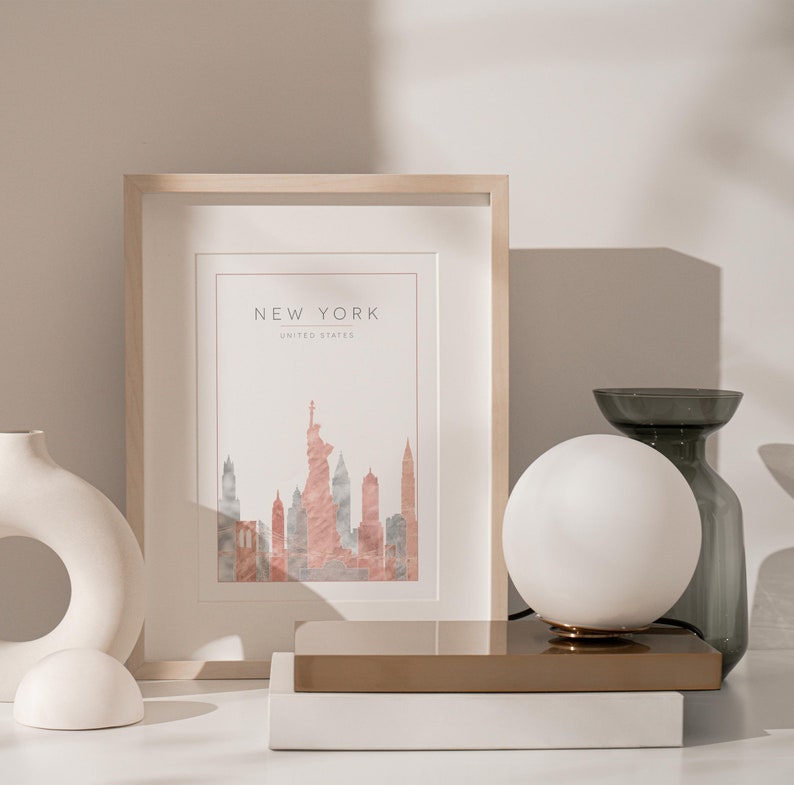May include: A framed print of a watercolor illustration of the New York City skyline, including the Statue of Liberty, with the text "NEW YORK, UNITED STATES" at the top. The print is on a white background and is framed in a light wood frame. The print is sitting on a white surface next to a white ceramic vase and a white globe lamp on a gold metal base.