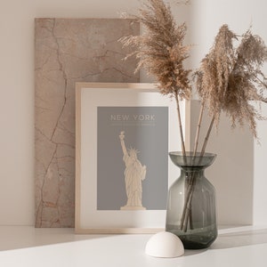 May include: A framed print with a grey background and a line drawing of the Statue of Liberty. The print says "New York, United States of America". The print is sitting on a white surface with a vase of dried pampas grass.