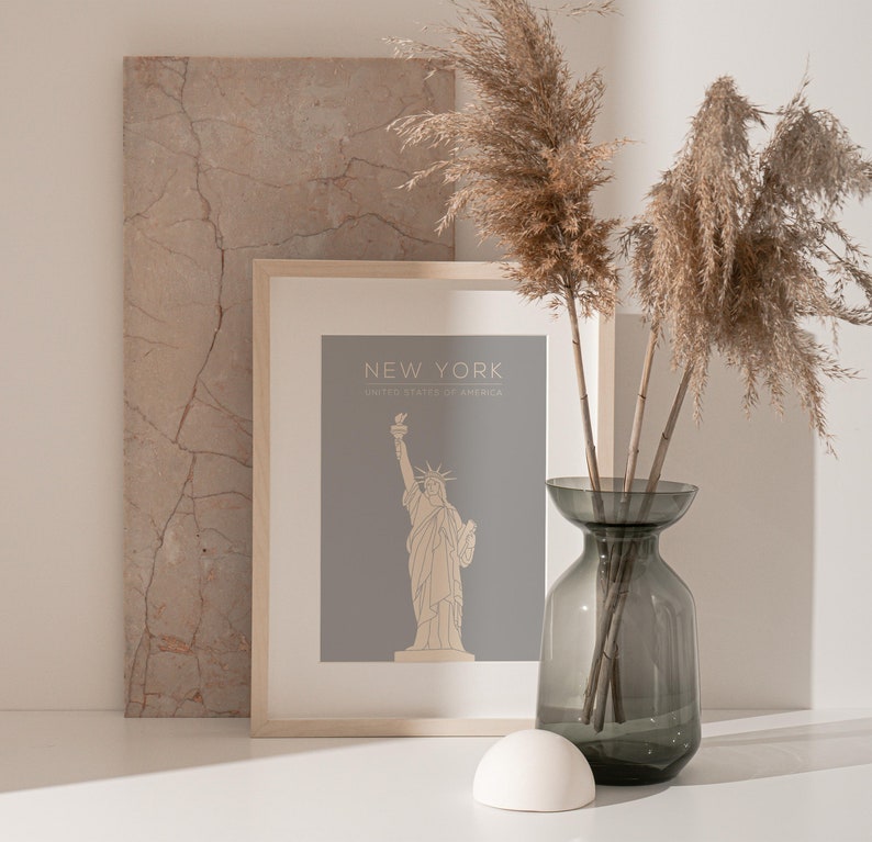 May include: A framed print with a grey background and a line drawing of the Statue of Liberty. The print says "New York, United States of America". The print is sitting on a white surface with a vase of dried pampas grass.