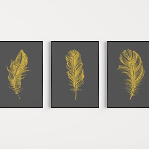 May include: Three framed prints with a dark gray background and a single gold feather illustration on each print.
