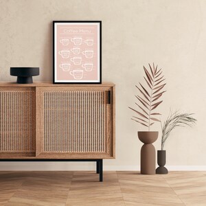 May include: A framed print with a pink background and white text that reads "Coffee Menu". The print features illustrations of different types of coffee cups, with the names of each type of coffee listed below the cups. The print is hanging on a wall above a wooden cabinet with a rattan front.