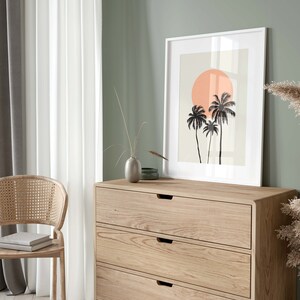 May include: A white framed print with a minimalist design of three palm trees and a large orange sun on a light beige background. The print is hanging on a light green wall above a wooden dresser with three drawers.