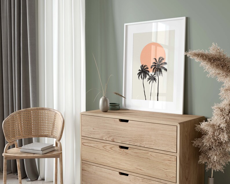 May include: A white framed print with a minimalist design of three palm trees and a large orange sun on a light beige background. The print is hanging on a light green wall above a wooden dresser with three drawers.