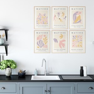 May include: Six framed prints of Matisse's "Paper Cutouts" artwork. Each print features a different cutout design in shades of pink, purple, and yellow. The prints are arranged in a grid pattern on a white wall above a kitchen sink.