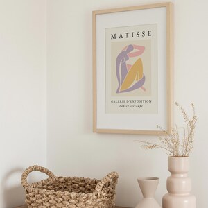 May include: A framed print of a Matisse artwork featuring a colorful abstract figure with the text "MATISSE" and "GALERIE D'EXPOSITION Papier Decoupe" in a light wood frame. The print is displayed on a wooden shelf with a woven basket and two ceramic vases.
