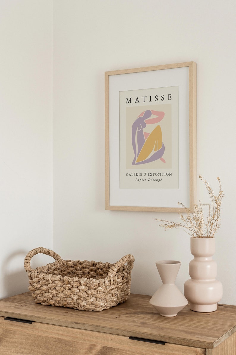 May include: A framed print of a Matisse artwork featuring a colorful abstract figure with the text "MATISSE" and "GALERIE D'EXPOSITION Papier Decoupe" in a light wood frame. The print is displayed on a wooden shelf with a woven basket and two ceramic vases.