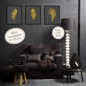 May include: Three framed prints of gold feathers on a dark gray background, hanging on a dark gray wall. A black armchair with black and white pillows sits in front of the prints. A black lamp with a white base stands next to the armchair. A black side table with a black teapot and a black cup sits next to the lamp. The text "Also available in store" and "Save over 30%" is visible in the image.