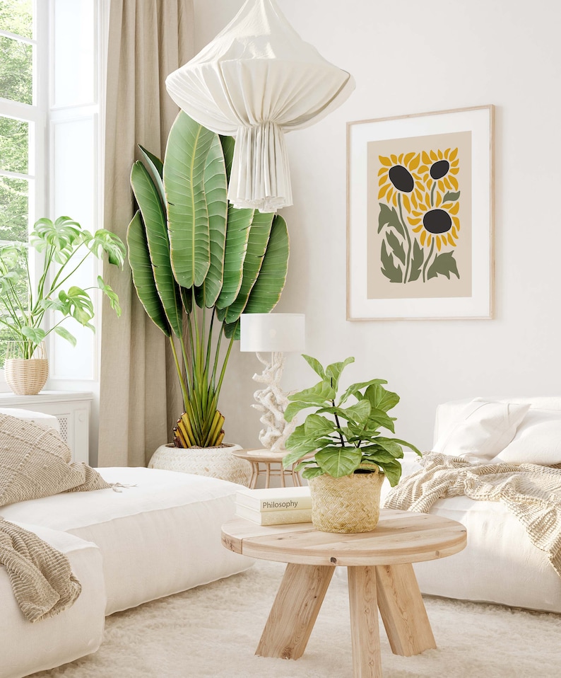 May include: A living room interior with a white couch, a coffee table, a potted plant, and a framed print of stylized sunflowers. The print has a beige background with green leaves and yellow sunflowers.