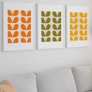 May include: Three framed prints with a simple abstract design of stylized leaves in orange, green, and yellow. The prints are hung on a white wall above a gray couch.