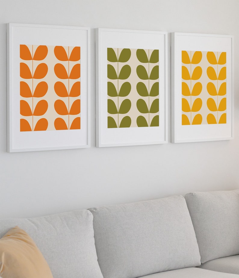 May include: Three framed prints with a simple abstract design of stylized leaves in orange, green, and yellow. The prints are hung on a white wall above a gray couch.
