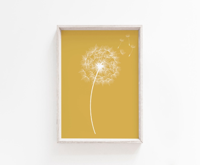 Op de afbeelding: Een ingelijste print van een witte paardenbloem met zaden die in de wind waaien op een gele achtergrond.