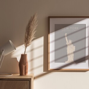 May include: A framed print featuring a minimalist line drawing of the Statue of Liberty against a light grey background. The text "NEW YORK" and "UNITED STATES OF AMERICA" is printed beneath the image. The print is hanging on a beige wall with a white lamp and a vase containing dried pampas grass on a wooden table in front of it.
