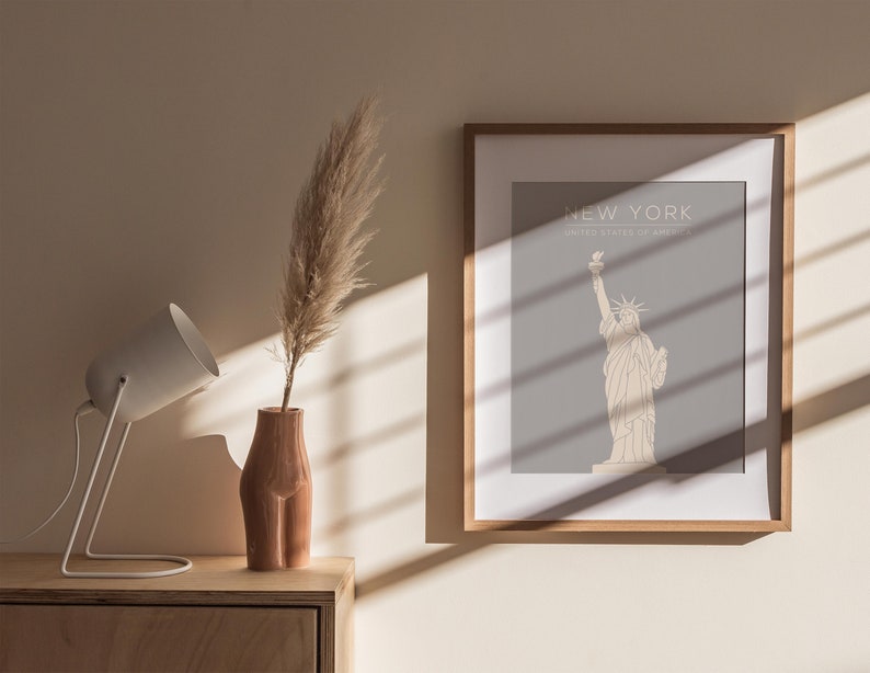 May include: A framed print featuring a minimalist line drawing of the Statue of Liberty against a light grey background. The text "NEW YORK" and "UNITED STATES OF AMERICA" is printed beneath the image. The print is hanging on a beige wall with a white lamp and a vase containing dried pampas grass on a wooden table in front of it.