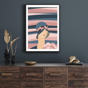 May include: A framed art print depicting a woman in a blue cloche hat with a pink bow. The background features horizontal stripes in pink and blue hues. The artwork is displayed on a dark wood chest of drawers.