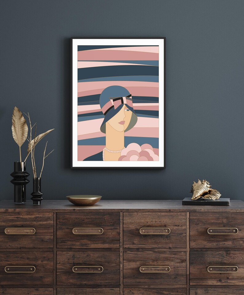 May include: A framed art print depicting a woman in a blue cloche hat with a pink bow. The background features horizontal stripes in pink and blue hues. The artwork is displayed on a dark wood chest of drawers.