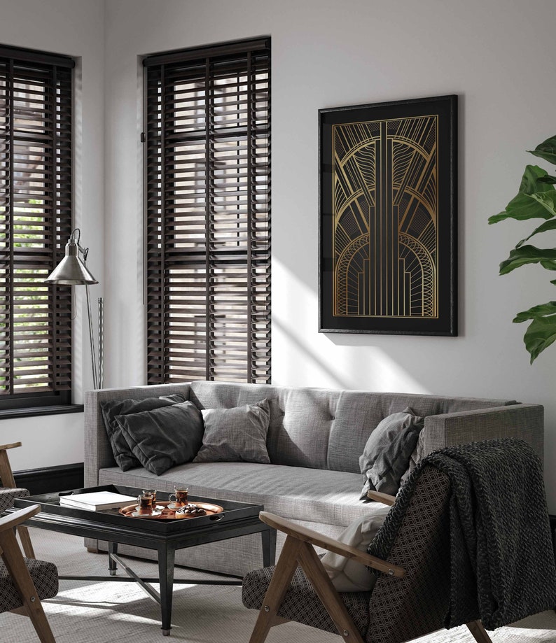 May include: A gray sofa with black throw pillows sits in a living room with brown wood blinds, a coffee table with a tray of glasses, and a black framed print with a gold geometric design.