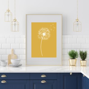 Op de afbeelding: Een ingelijste print van een witte paardenbloem met zaden die in de wind waaien tegen een gele achtergrond. De print hangt aan een witte betegelde muur boven een blauw keukenkastje.