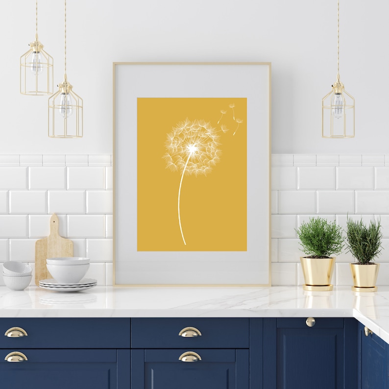 Op de afbeelding: Een ingelijste print van een witte paardenbloem met zaden die in de wind waaien tegen een gele achtergrond. De print hangt aan een witte betegelde muur boven een blauw keukenkastje.