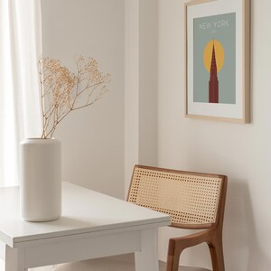 May include: A white table with a white vase filled with dried flowers sits next to a brown wicker chair. A framed print with the text "NEW YORK, USA" and a stylized image of a skyscraper is hanging on the wall.