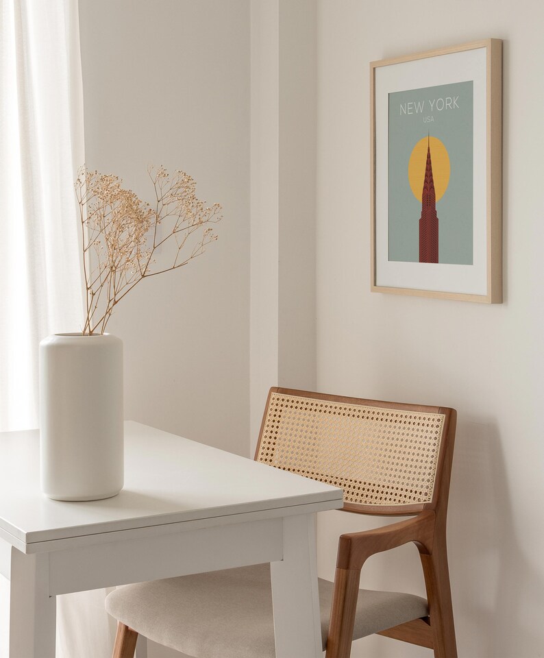 May include: A white table with a white vase filled with dried flowers sits next to a brown wicker chair. A framed print with the text "NEW YORK, USA" and a stylized image of a skyscraper is hanging on the wall.