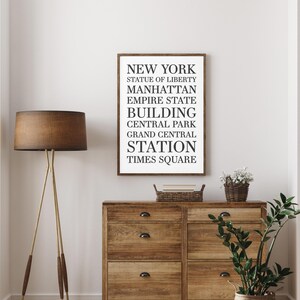 May include: A framed black and white print with the text "NEW YORK" and a list of iconic New York City landmarks, including the Statue of Liberty, Manhattan, Empire State Building, Central Park, Grand Central Station, and Times Square. The print is hanging on a white wall above a wooden dresser.