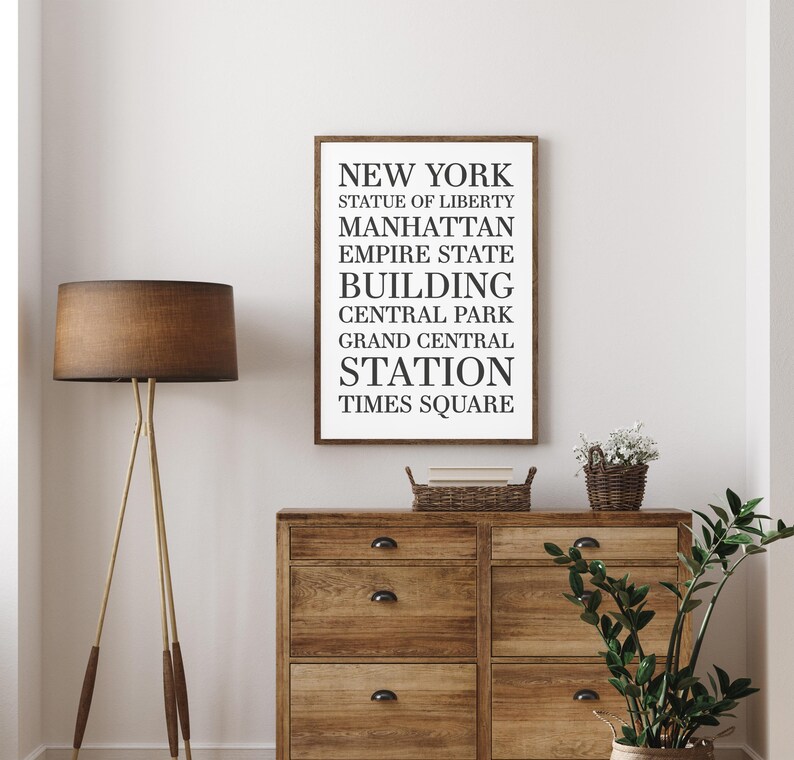 May include: A framed black and white print with the text "NEW YORK" and a list of iconic New York City landmarks, including the Statue of Liberty, Manhattan, Empire State Building, Central Park, Grand Central Station, and Times Square. The print is hanging on a white wall above a wooden dresser.
