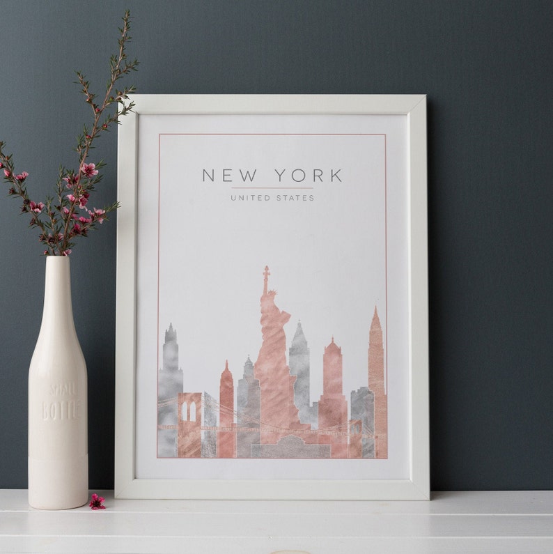May include: A white framed print with a watercolor illustration of the New York City skyline in shades of pink and gray. The text "NEW YORK" and "UNITED STATES" is printed in gray at the top of the print.
