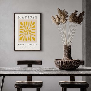 以下が含まれることがあります： 黄色い抽象的なデザインと「MATISSE MUS&Eacute;E D'ORSAY Rue de la L&eacute;gion d'honneur」というテキストが書かれた、マティスの作品を額装したプリント。プリントは灰色の壁にかけられており、その下には木製のテーブルと2つの椅子、そして乾燥したパンパスグラスの入った籐の壺が置かれている。