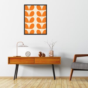 May include: A mid-century modern living room with a wooden console table, a lamp, a vase with branches, and a framed print of orange leaf designs on a white background. A gray armchair sits to the right of the table.