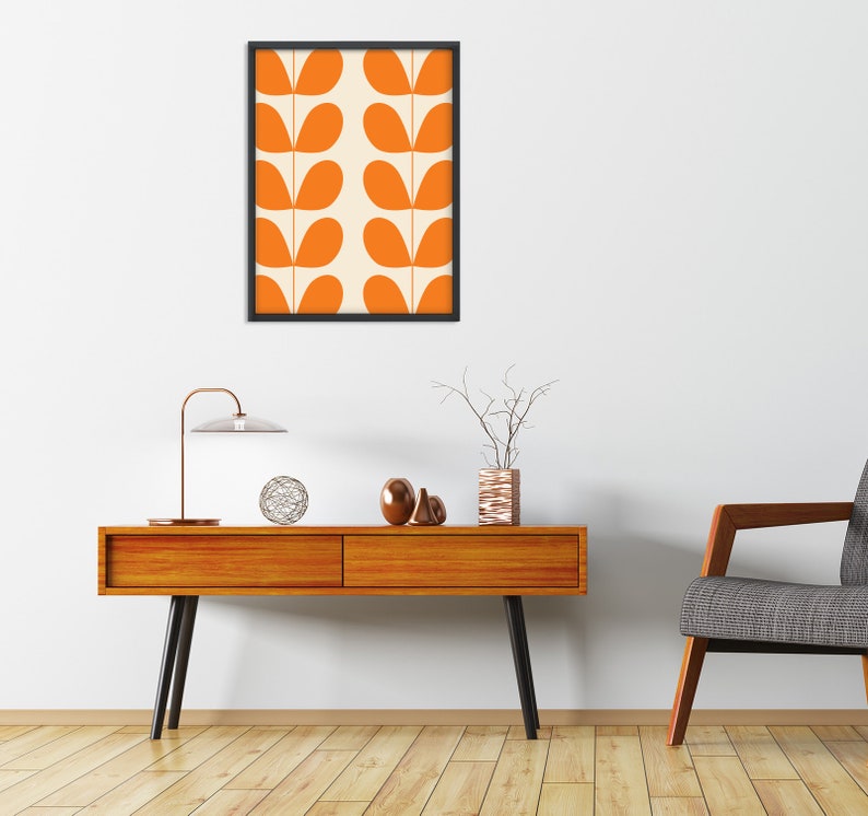 May include: A mid-century modern living room with a wooden console table, a lamp, a vase with branches, and a framed print of orange leaf designs on a white background. A gray armchair sits to the right of the table.