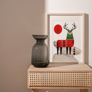 May include: A grey glass vase with a wide mouth sits on a wooden table with a woven drawer. Behind the vase is a framed print of a stylised deer wearing a red and green sweater.