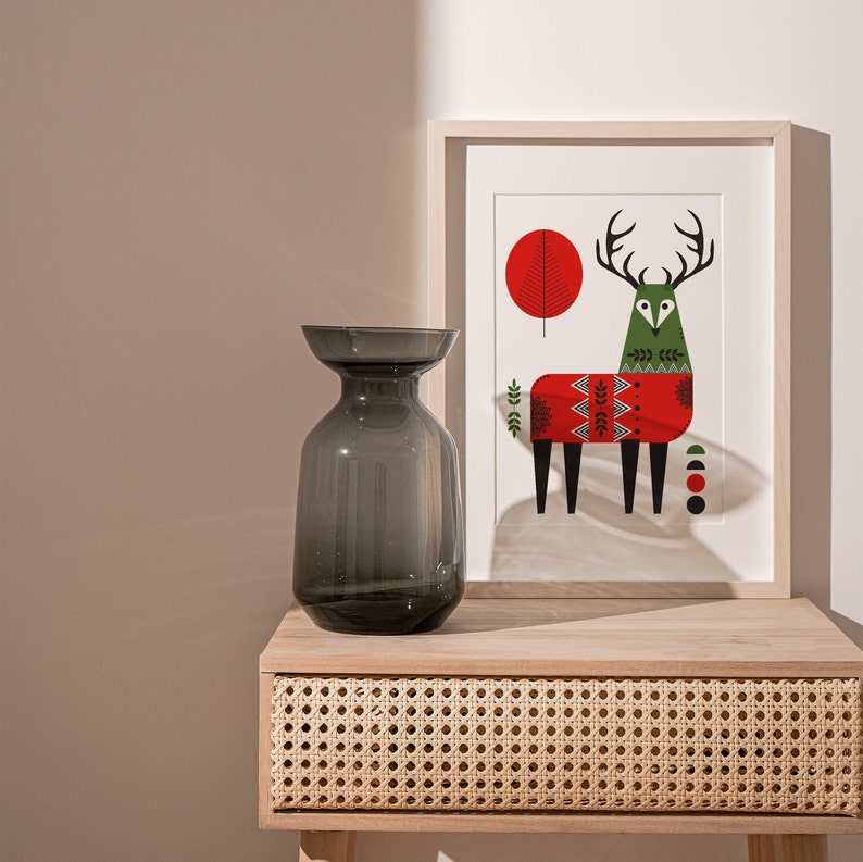 May include: A grey glass vase with a wide mouth sits on a wooden table with a woven drawer. Behind the vase is a framed print of a stylised deer wearing a red and green sweater.