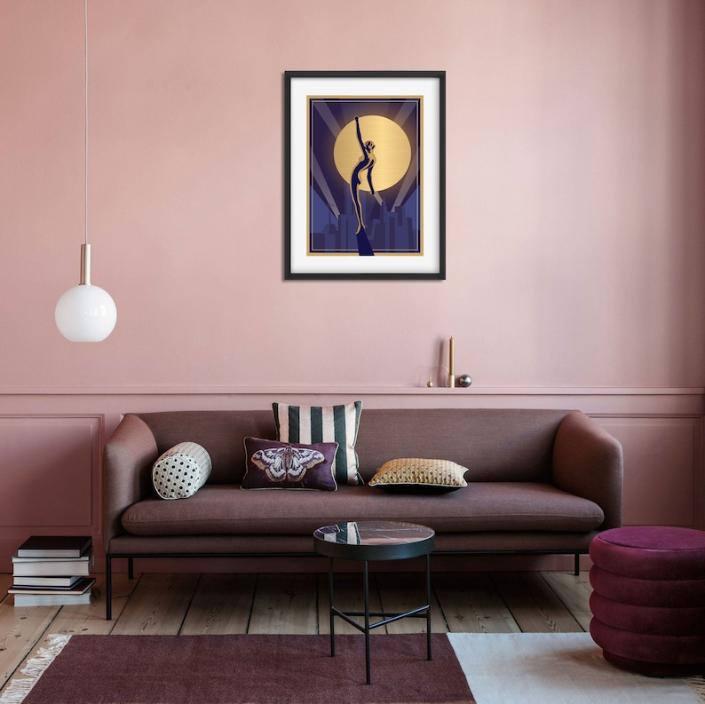 May include: A framed art print with a gold silhouette of a woman standing on a platform in front of a city skyline. The woman is reaching up towards a large, golden moon.