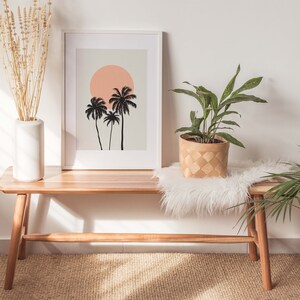 May include: A wooden bench with a white faux fur throw and a potted plant with green leaves. A framed print with a silhouette of palm trees and a pink sun hangs on the wall above the bench.