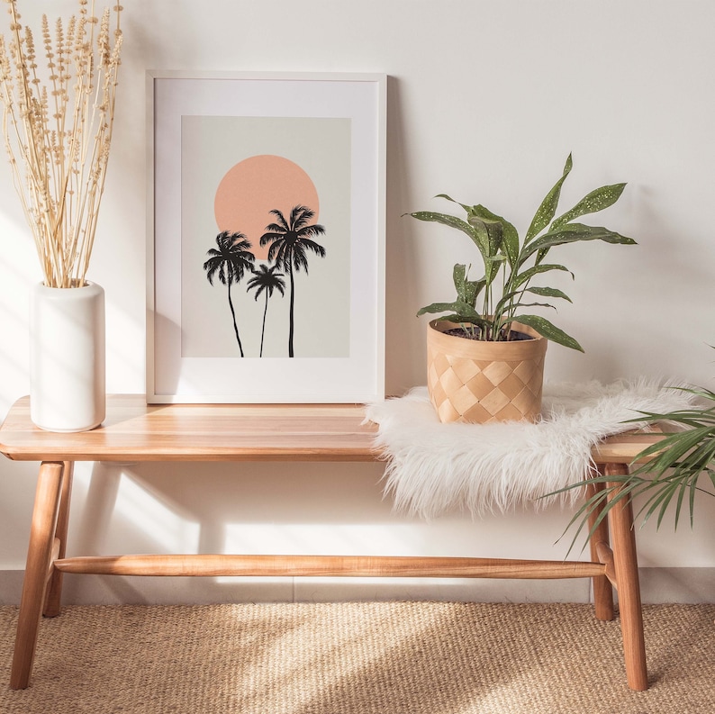 May include: A wooden bench with a white faux fur throw and a potted plant with green leaves. A framed print with a silhouette of palm trees and a pink sun hangs on the wall above the bench.