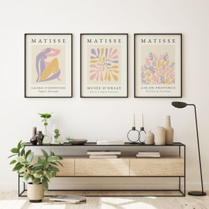 May include: Three framed art prints with a white background and black frames. The prints feature abstract designs in pink, purple, and yellow. The text on the prints reads "MATISSE", "GALERIE D'EXPOSITION Papier D&eacute;coup&eacute;", "MUS&Eacute;E D'ORSAY Rue de la L&eacute;gion d'honneur", and "AIX-EN-PROVENCE Pavillon de Vend&ocirc;me".