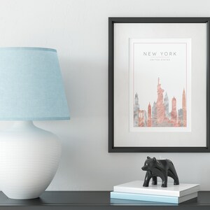 May include: A black framed print with a watercolor illustration of the New York City skyline in shades of pink and gray. The text "NEW YORK" and "UNITED STATES" is printed below the illustration. The print is displayed on a white wall above a black table with a white lamp and a black bear candle holder.