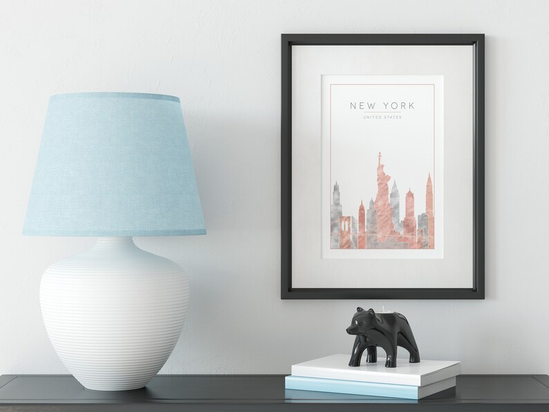 May include: A black framed print with a watercolor illustration of the New York City skyline in shades of pink and gray. The text "NEW YORK" and "UNITED STATES" is printed below the illustration. The print is displayed on a white wall above a black table with a white lamp and a black bear candle holder.