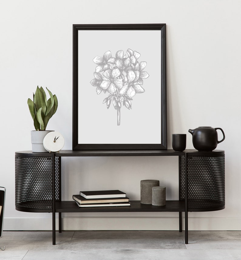 May include: A black metal console table with two shelves. The top shelf has a black framed print of a gray floral illustration. The bottom shelf has a stack of books and two gray cylindrical containers.