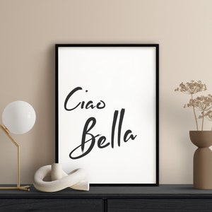 May include: Black and white framed print with the text "Ciao Bella" in a handwritten style. The print is displayed on a black shelf with a white ceramic sculpture, a gold lamp, and a brown vase with dried flowers.